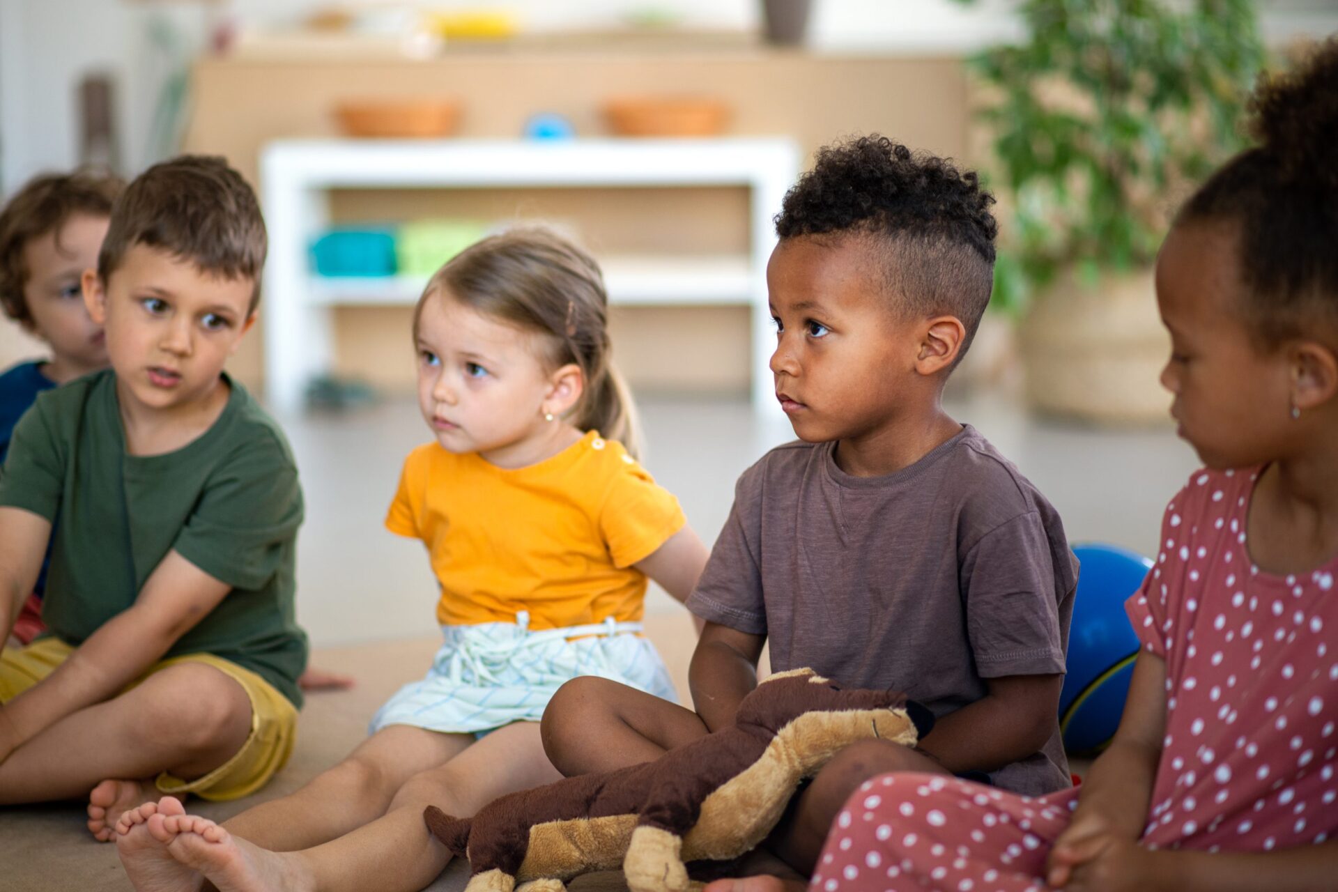 Group of small nursery school children sitting on floor indoors in classroom, listening to teacher. A group of small nursery school children sitting on floor indoors in classroom, listening to teacher.